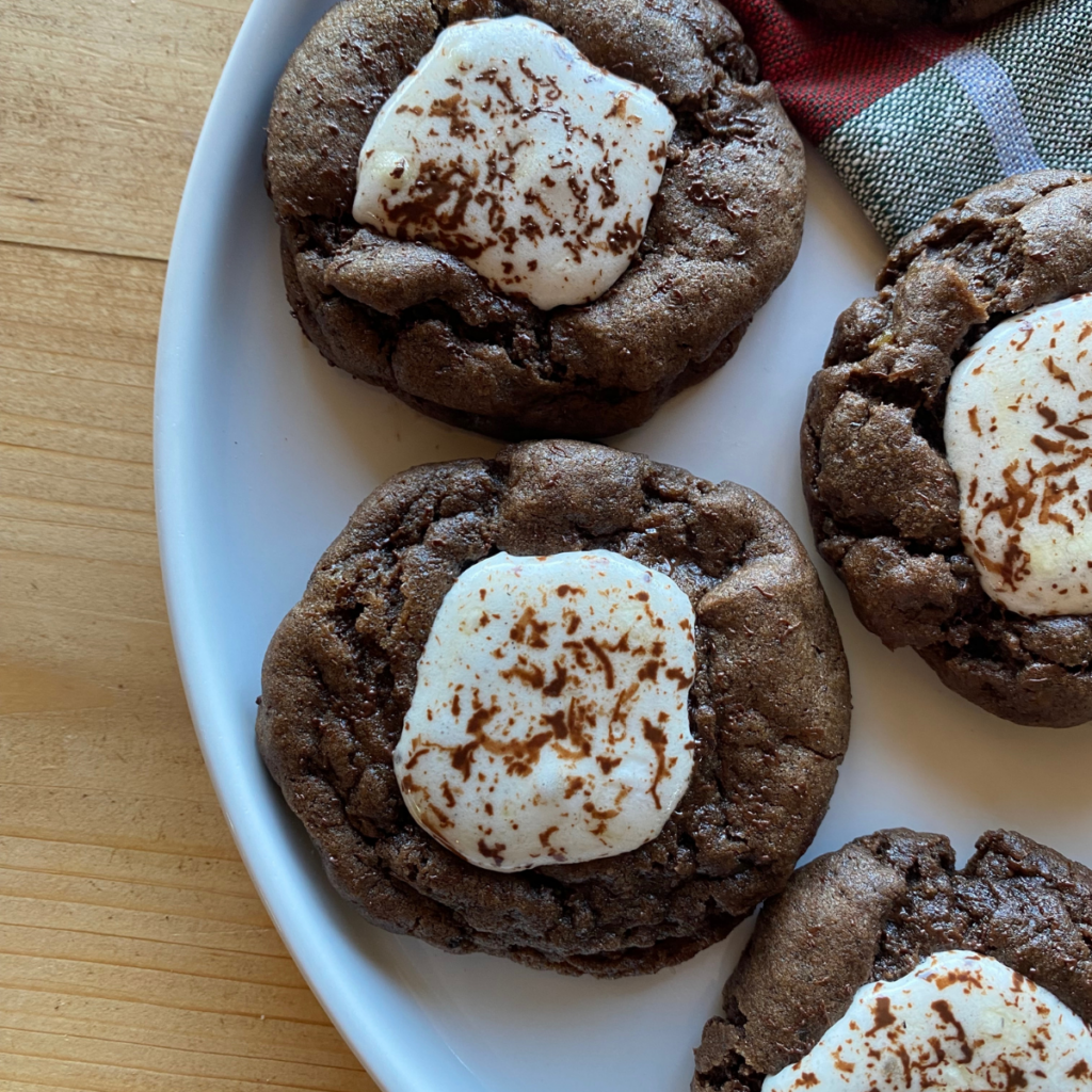 Soft and chewy hot chocolate cookies topped with toasted marshmallows on a white plate — a cozy winter dessert perfect for holiday baking.
