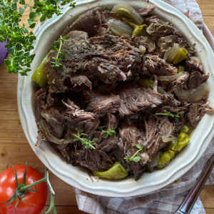 Shredded Italian beef sitting in a bowl, a tomato and greenery are in the background