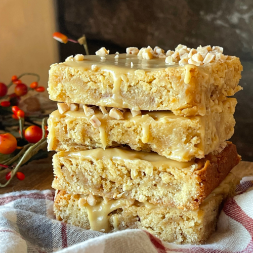 Stack of gooey sourdough blondies drizzled with maple toffee glaze and topped with chopped toffee bits, showing their chewy, golden texture on a rustic fall background.”