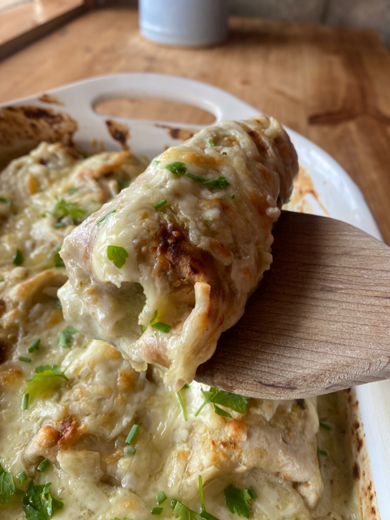 a close up view of salsa verde chicken enchiladas. One enchilada is lifted from the dish by a wooden spatula