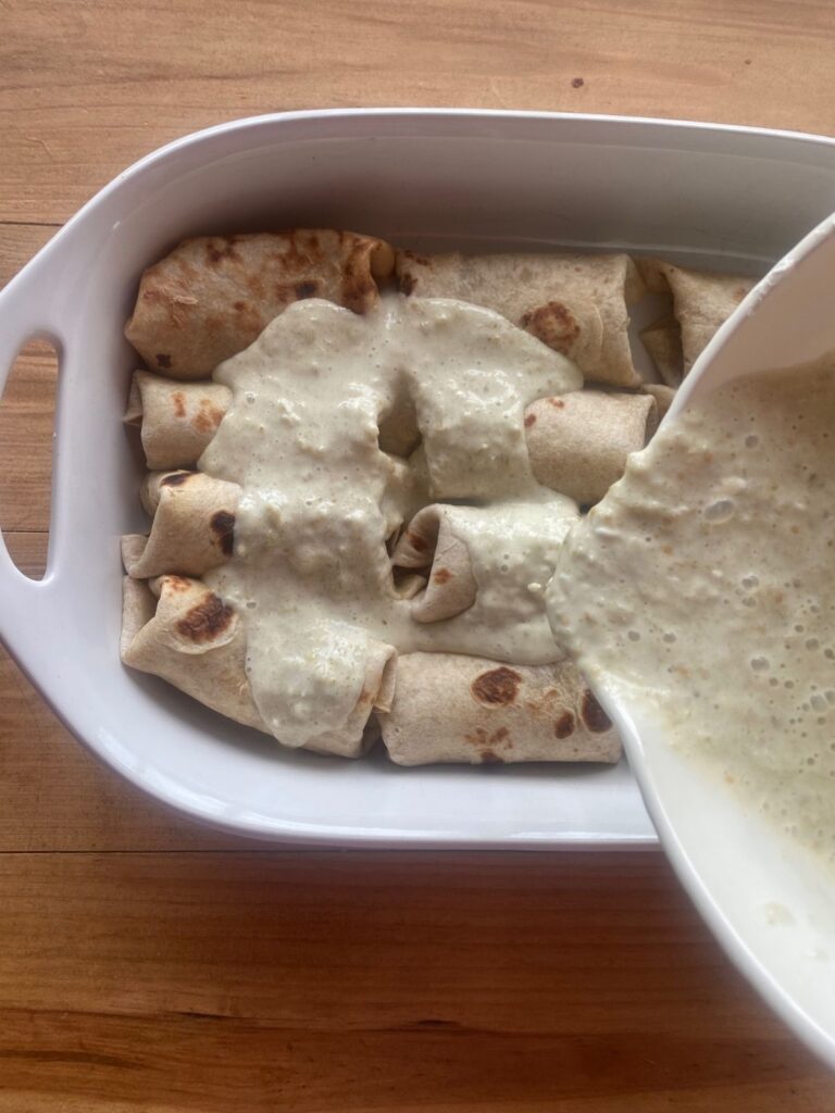 rolled salsa verde chicken enchiladas assembled in a white baking dish. A creamy sauce is being poured over the top