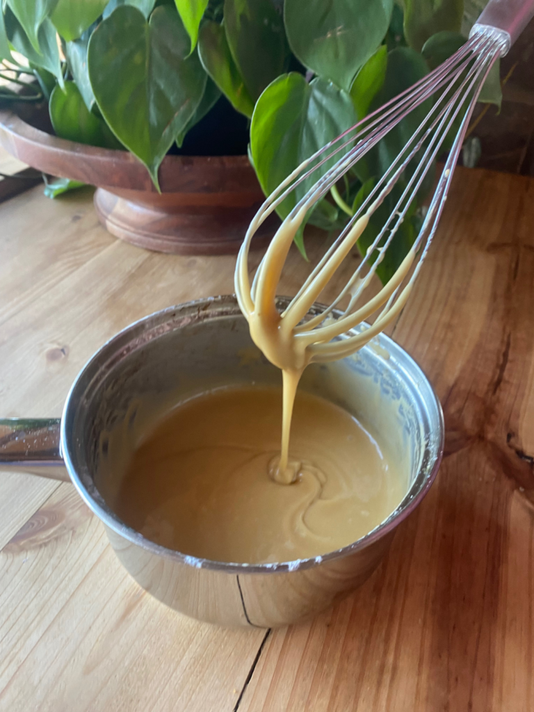 A pan of maple glaze sitting on a wooden countertop. A whisk is hanging over the top, with glaze drizzling down from the whisk into the pan