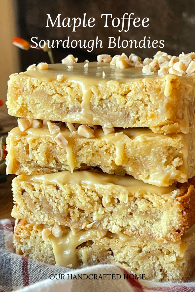 Stack of gooey sourdough blondies drizzled with maple toffee glaze and topped with chopped toffee bits, showing their chewy, golden texture on a rustic fall background. Text overlay reads "Maple Toffee Sourdough Blondies"