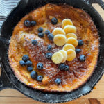 a sourdough Dutch baby pancake baked in a cast iron skillet and topped with blueberries, sliced bananas, and a drizzle of maple syrup