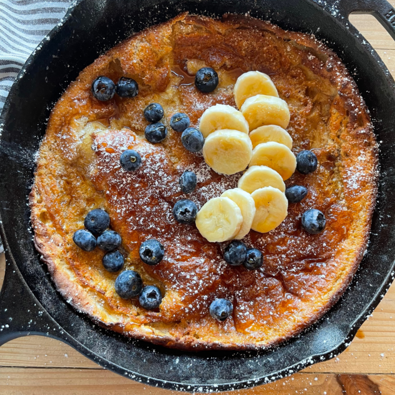 a sourdough Dutch baby pancake baked in a cast iron skillet and topped with blueberries, sliced bananas, and a drizzle of maple syrup