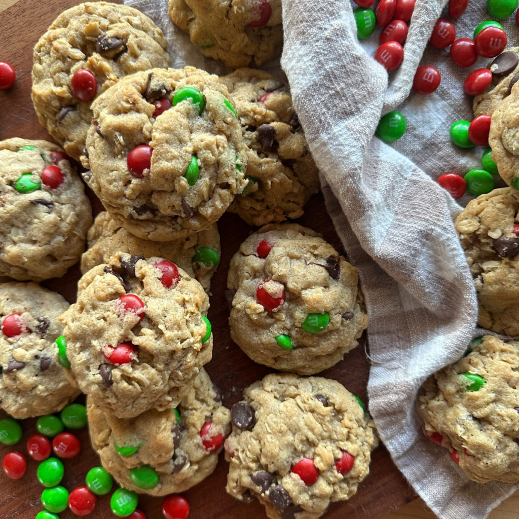Christmas monster cookies piled on a wooden board, packed with oats, chocolate chips, and red and green M&M candies for a festive holiday treat.