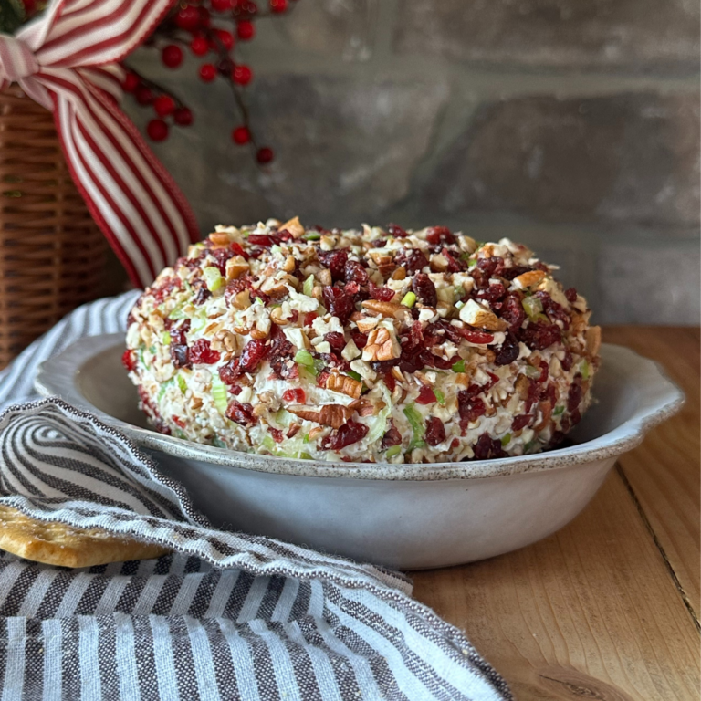 A cheese ball coated with chopped pecans and cranberries, sitting in a gray dish. A striped towel is off to the side, along with a pile of crackers.