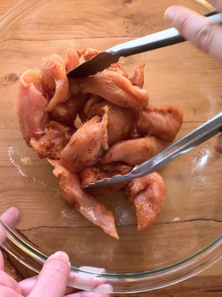 seasoned uncooked chicken breast being tossed together with tongs in a large glass bowl