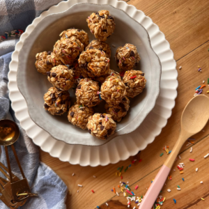 A bowl full of no bake protein balls sitting on a wooden countertop. Rainbow sprinkles are scattered around the bowl, while a wooden and pink spoon lays to the side