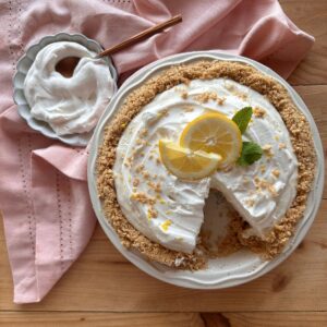a creamy no bake lemon pie is nestled in a graham cracker crust and garnished with lemon slices and mint sprigs. A light pink towel and a small bowl of whipped cream are off to the side.