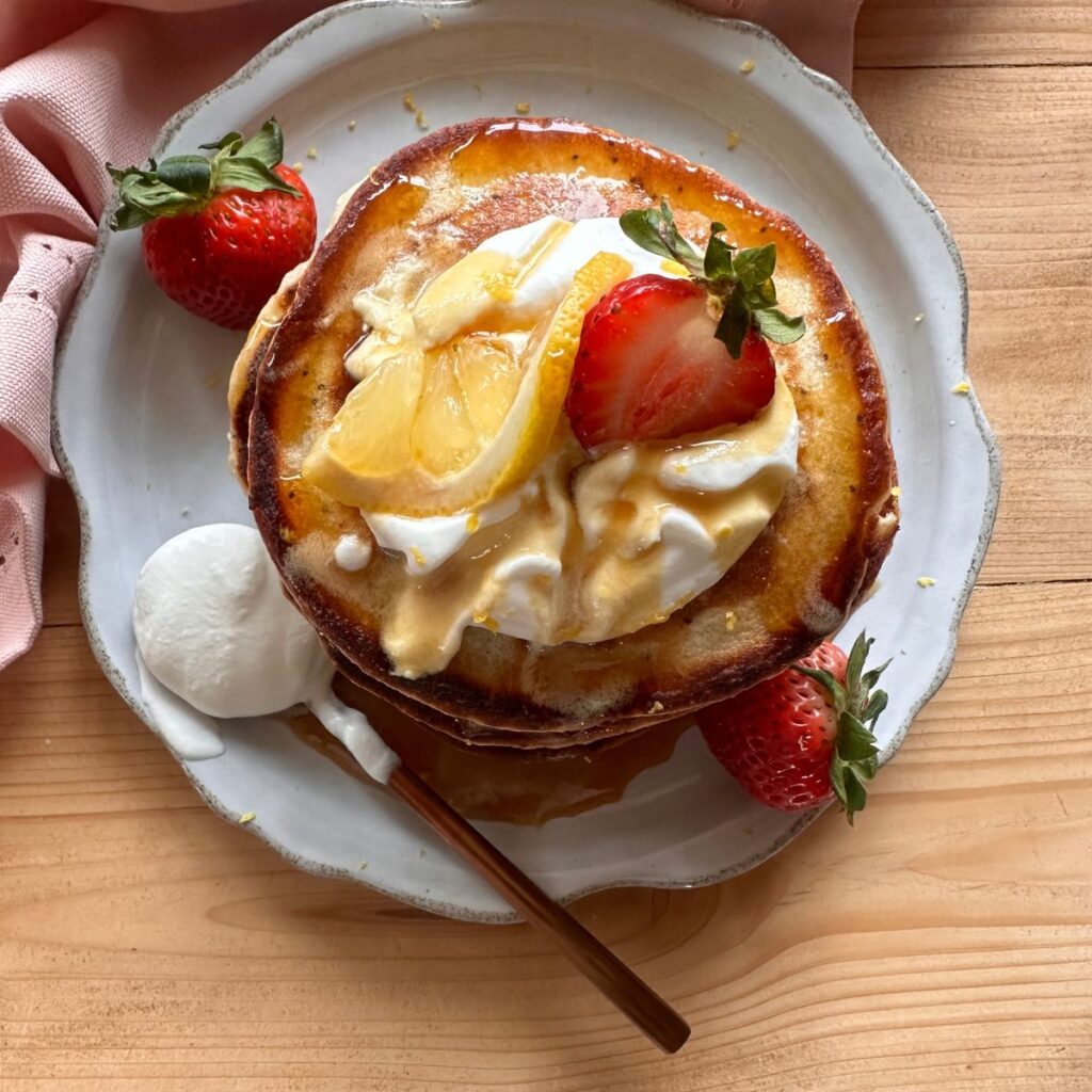 a stack of lemon poppyseed pancakes on a gray plate, topped with a dollop of whipped cream and garnished with strawberries and a lemon wedge.