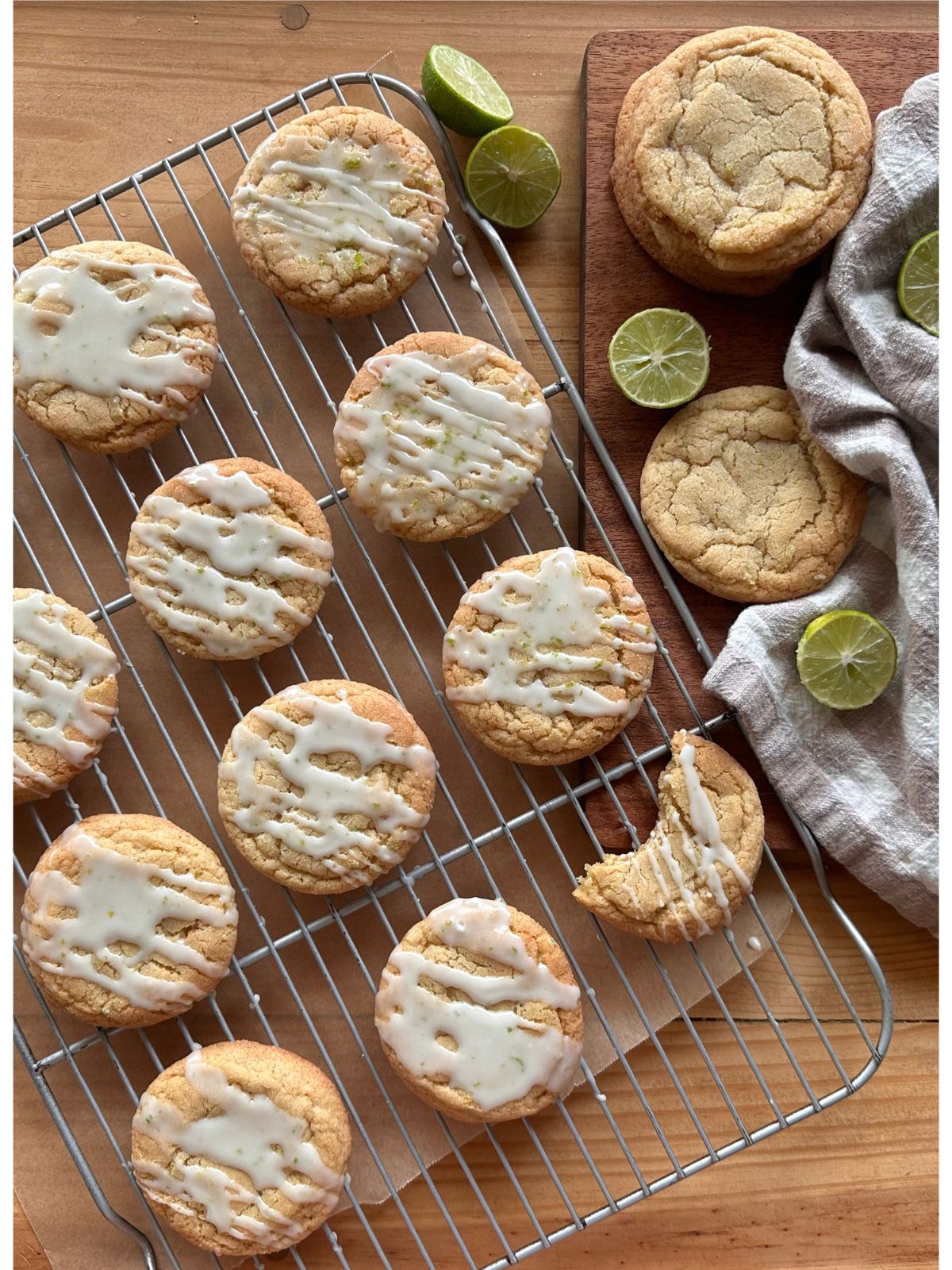 an arrangement of baked key lime cookies on a wire cooling rack, drizzled with glaze and garnished with lime zest. Sliced key limes are arranged around the cookies, while a checkered towel lies in the background.