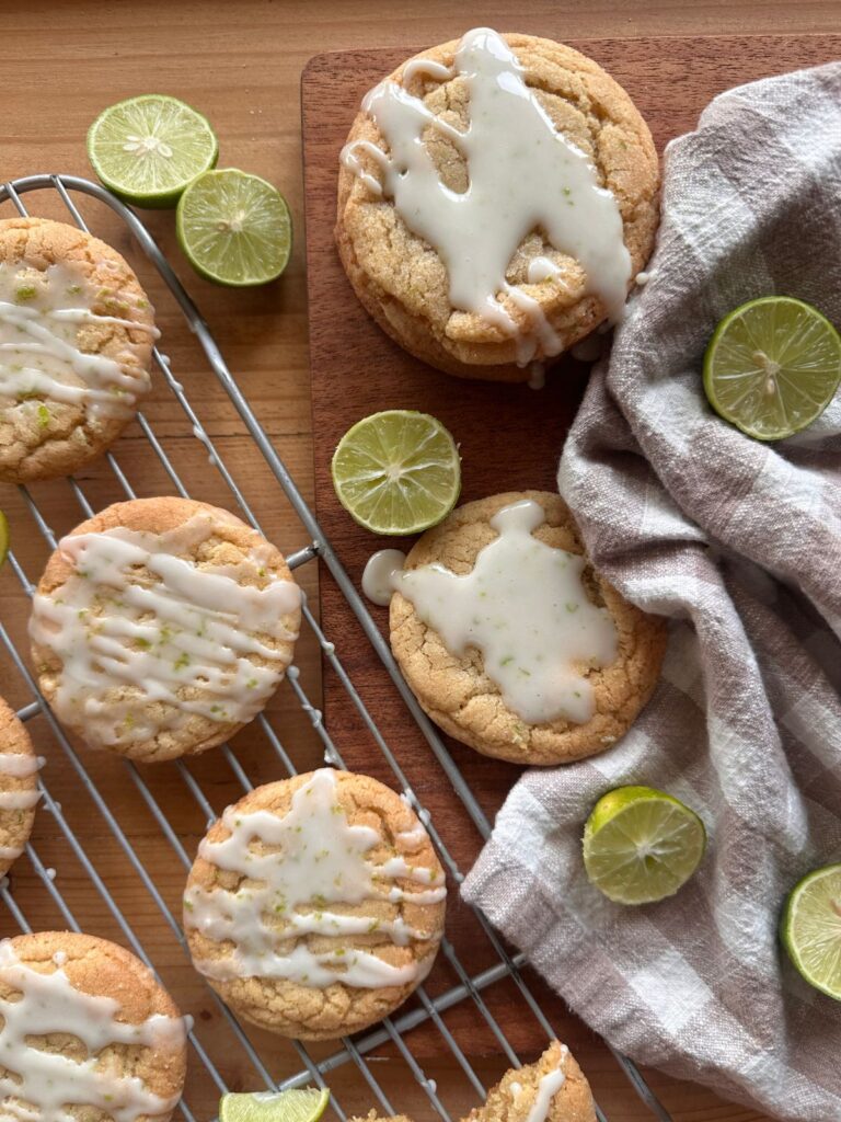 glazed key lime cookies arranged on a cooling rack. Sliced key limes are arranged around the cookies, along with a checkered kitchen towel