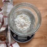 a jar of flour and water is set on top of a food scale. A checkered towel lays in the background, along with a spoon