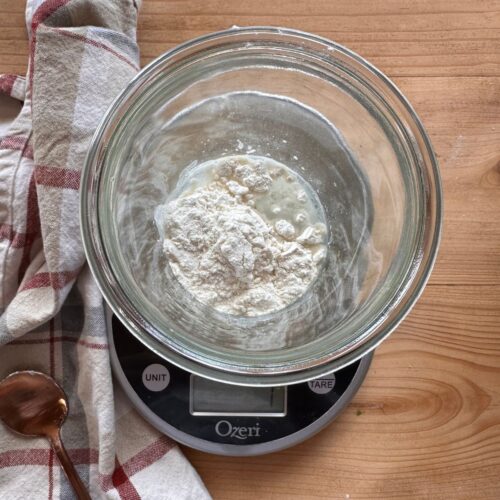 a jar of flour and water is set on top of a food scale. A checkered towel lays in the background, along with a spoon