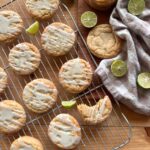an arrangement of baked key lime cookies on a wire cooling rack, drizzled with glaze and garnished with lime zest. Sliced key limes are arranged around the cookies, while a checkered towel lies in the background.