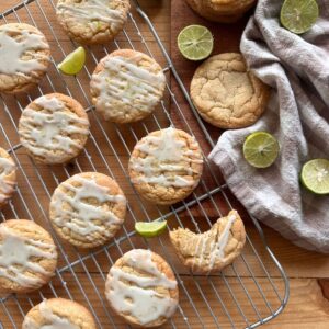 an arrangement of baked key lime cookies on a wire cooling rack, drizzled with glaze and garnished with lime zest. Sliced key limes are arranged around the cookies, while a checkered towel lies in the background.