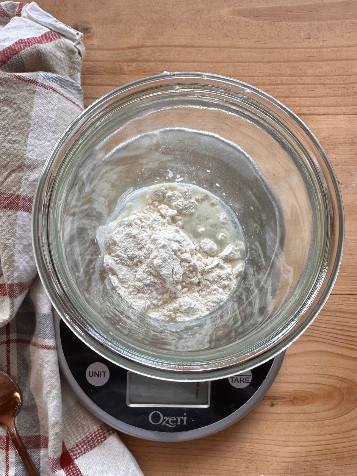 a jar of flour and water is set on top of a food scale to make homemade sourdough starter. A checkered towel lays in the background, along with a spoon