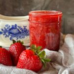a small jar of strawberry sauce sits on a checkered cloth, with whole strawberries around the outside of the jar. A blue and white dish sits in the background, behind the jar of strawberry sauce