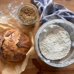 a freshly baked sourdough boule, a large glass jar of flour, and wheat berries in a glass jar, all arranged on a wooden countertop