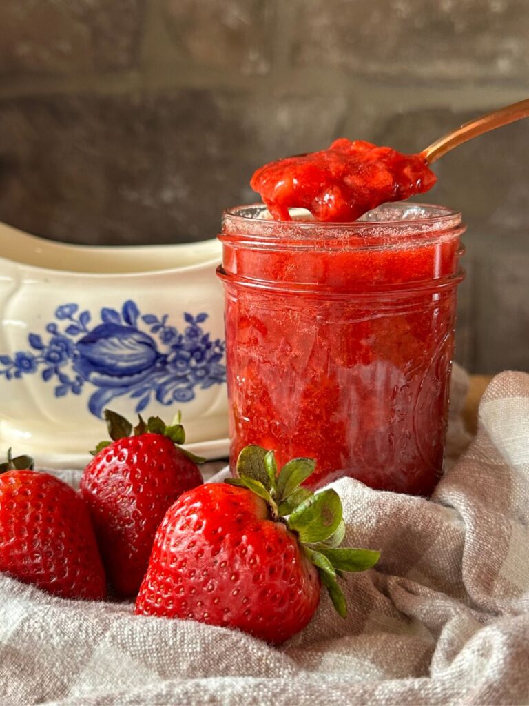 a jar of homemade strawberry sauce with a spoon hovering over the top of the jar with a spoonful of sauce.