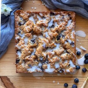 sourdough blueberry coffee cake laying on a wooden countertop, drizzled with vanilla icing. A blue cloth lays in the background, and blueberries are scattered around the edges of the cake