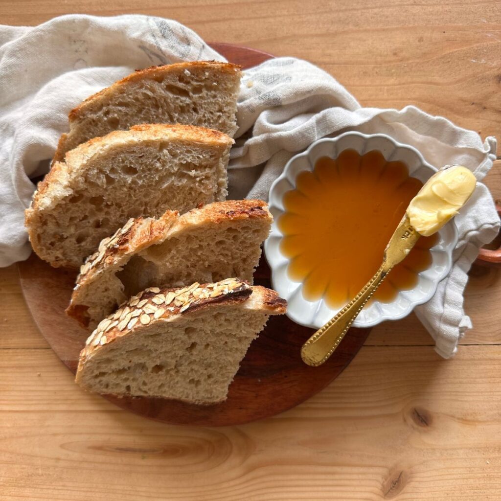 slices of sourdough oatmeal bread arranged on a wooden cutting board. A small dish of honey lays to the side, along with a butter knife