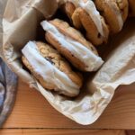 Sourdough chocolate chip cookies with vanilla ice cream sandwiched in the middle, aligned together in a small pan. The pan is lined with brown parchment paper, and is sitting on a wooden countertop.