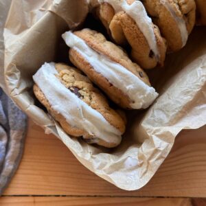 Sourdough chocolate chip cookies with vanilla ice cream sandwiched in the middle, aligned together in a small pan. The pan is lined with brown parchment paper, and is sitting on a wooden countertop.