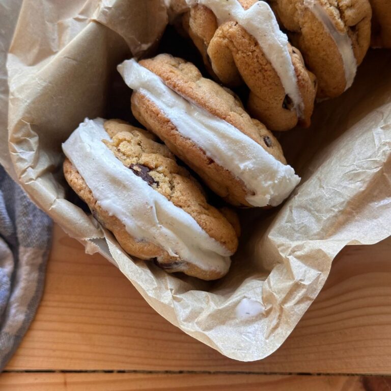 Sourdough chocolate chip cookies with vanilla ice cream sandwiched in the middle, aligned together in a small pan. The pan is lined with brown parchment paper, and is sitting on a wooden countertop.