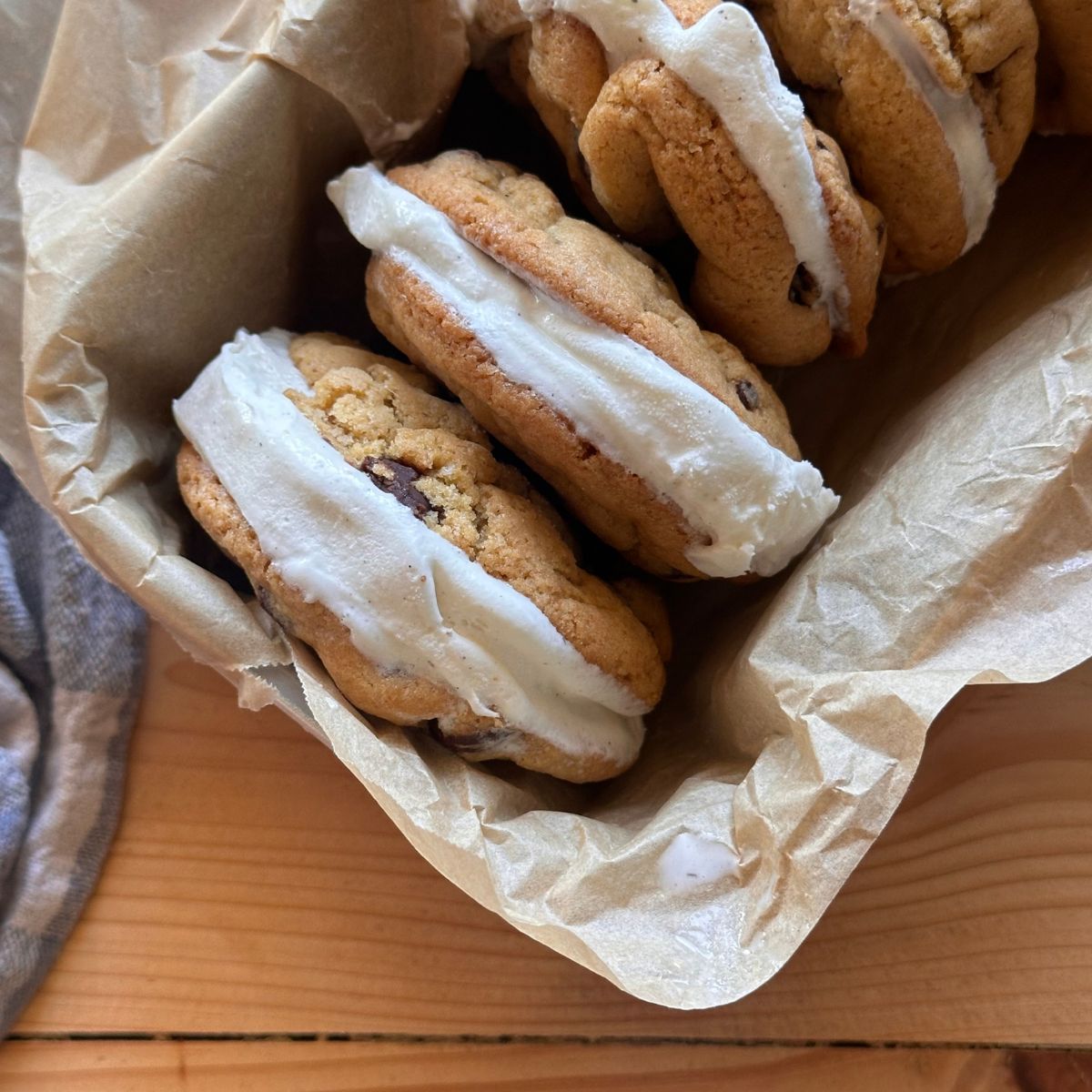 Sourdough chocolate chip cookies with vanilla ice cream sandwiched in the middle, aligned together in a small pan. The pan is lined with brown parchment paper, and is sitting on a wooden countertop.