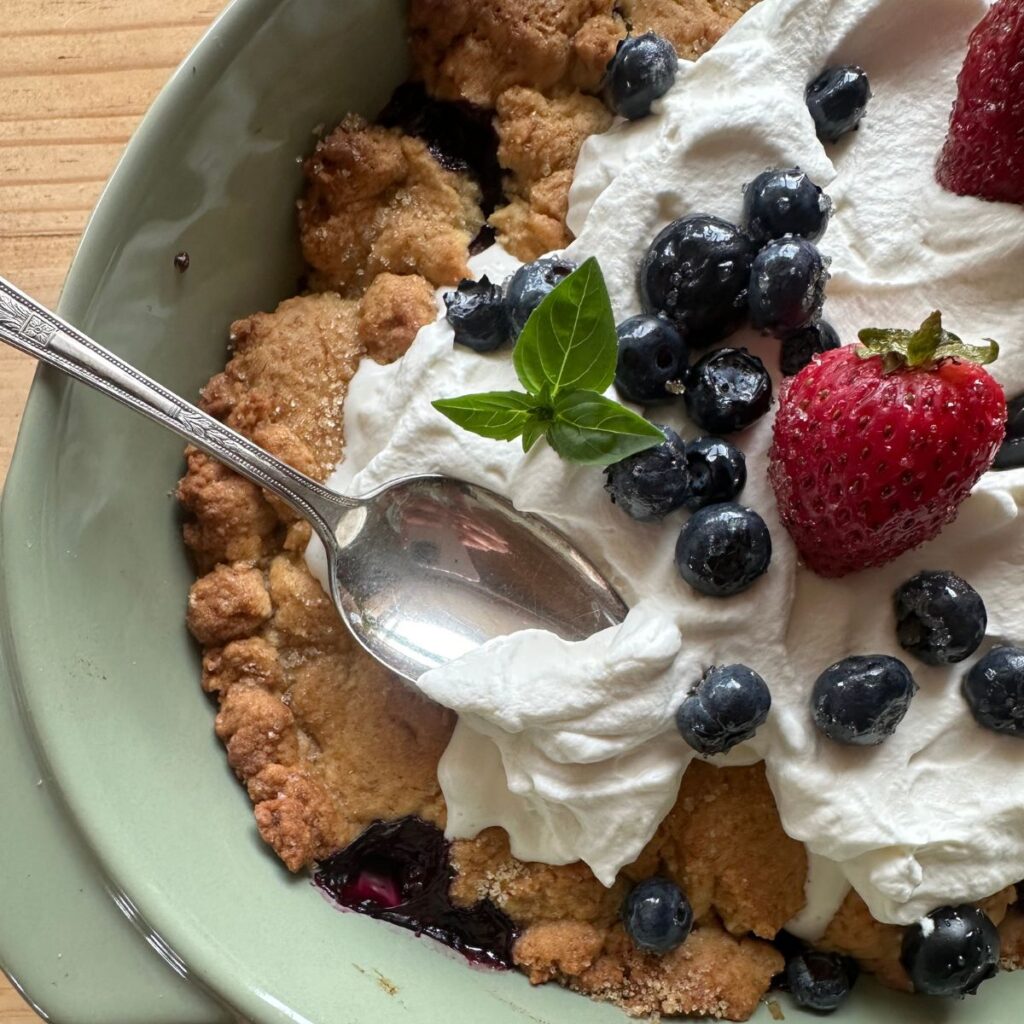 mixed berry cobbler in a green baking dish, topped with sourdough sugar cookie crust and swirls of whipped cream. Blueberries and strawberries are arranged over the top
