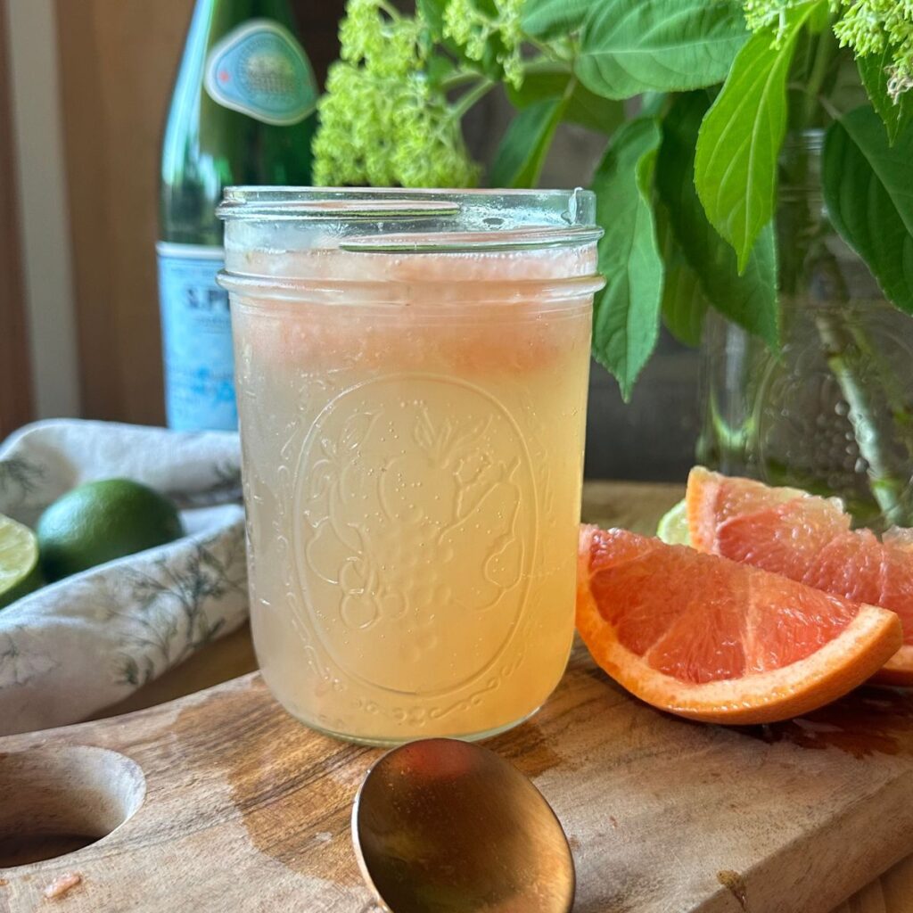 a close up view of a glass, filled with grapefruit & lime sparkling water. Sliced grapefruit lies to the side, along with a bottle of sparkling water in the background