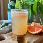 a close up view of a glass, filled with grapefruit & lime sparkling water. Sliced grapefruit lies to the side, along with a bottle of sparkling water in the background