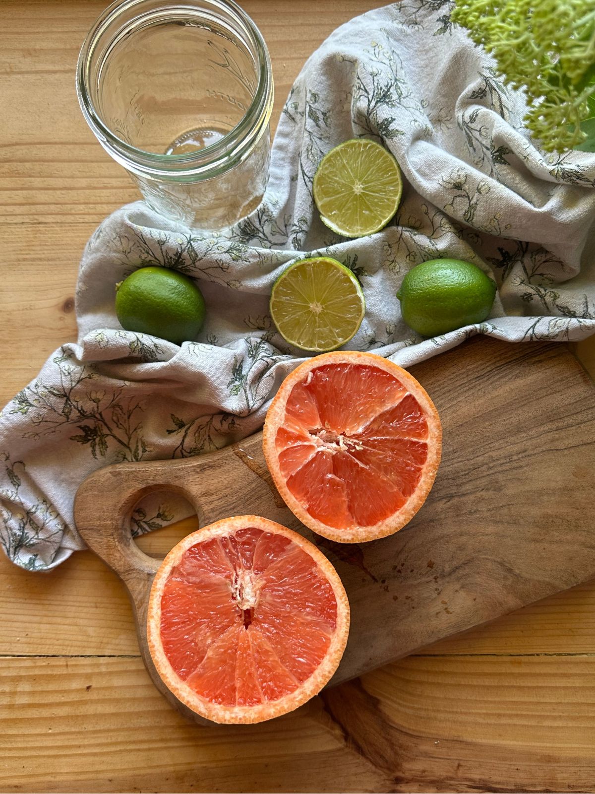sliced grapefruit and limes are arranged on a wooden countertop. A floral patterned kitchen towel lies in the background, along with a wooden cutting board.