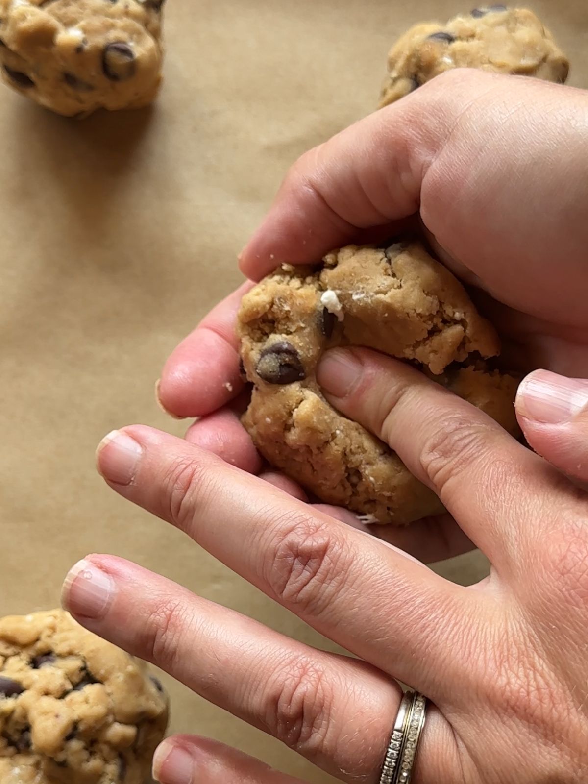 a hand holds a ball of cookie dough over a baking sheet, while a finger is gently pressed into the center of the dough, creating a small hole in the middle of the dough