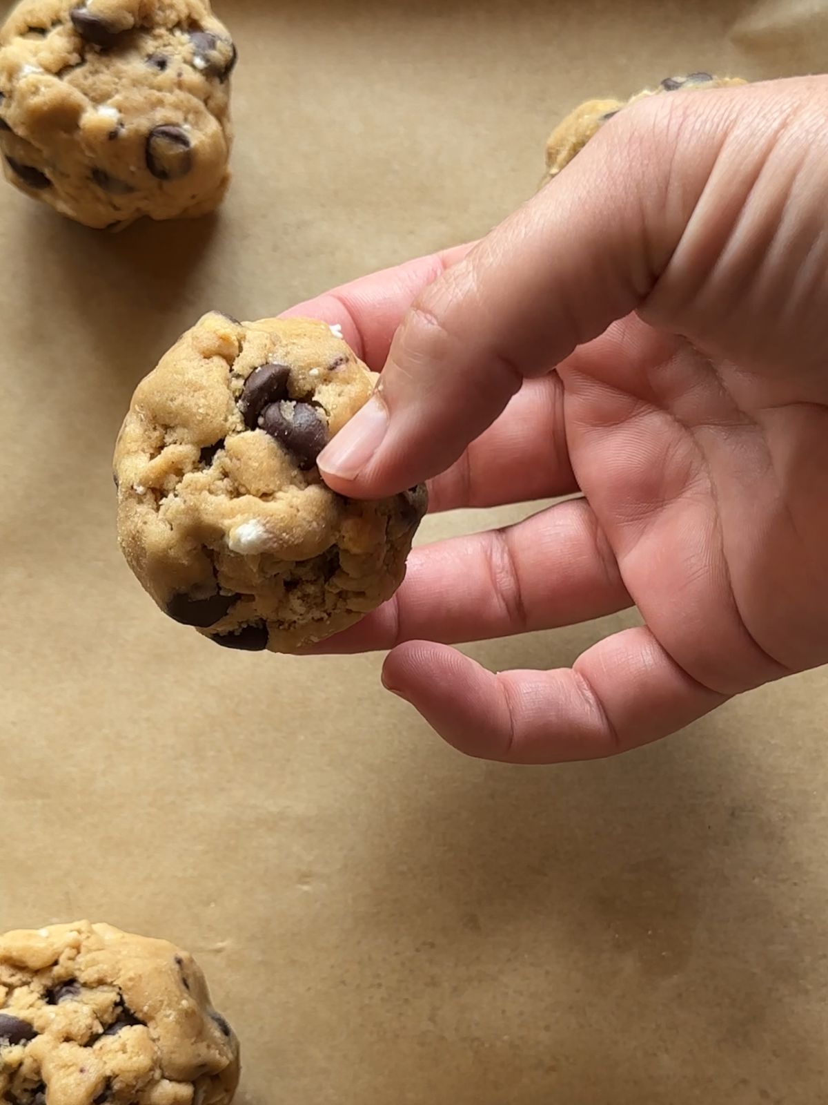 a hand holds a ball of cookie dough over a parchment paper lined baking sheet