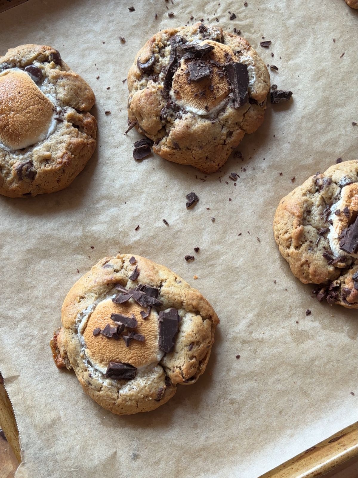 baked smores cookies on a parchment paper lined baking sheet. A toasty marshmallow is in the center of each cookie, and then garnished with chopped chocolate