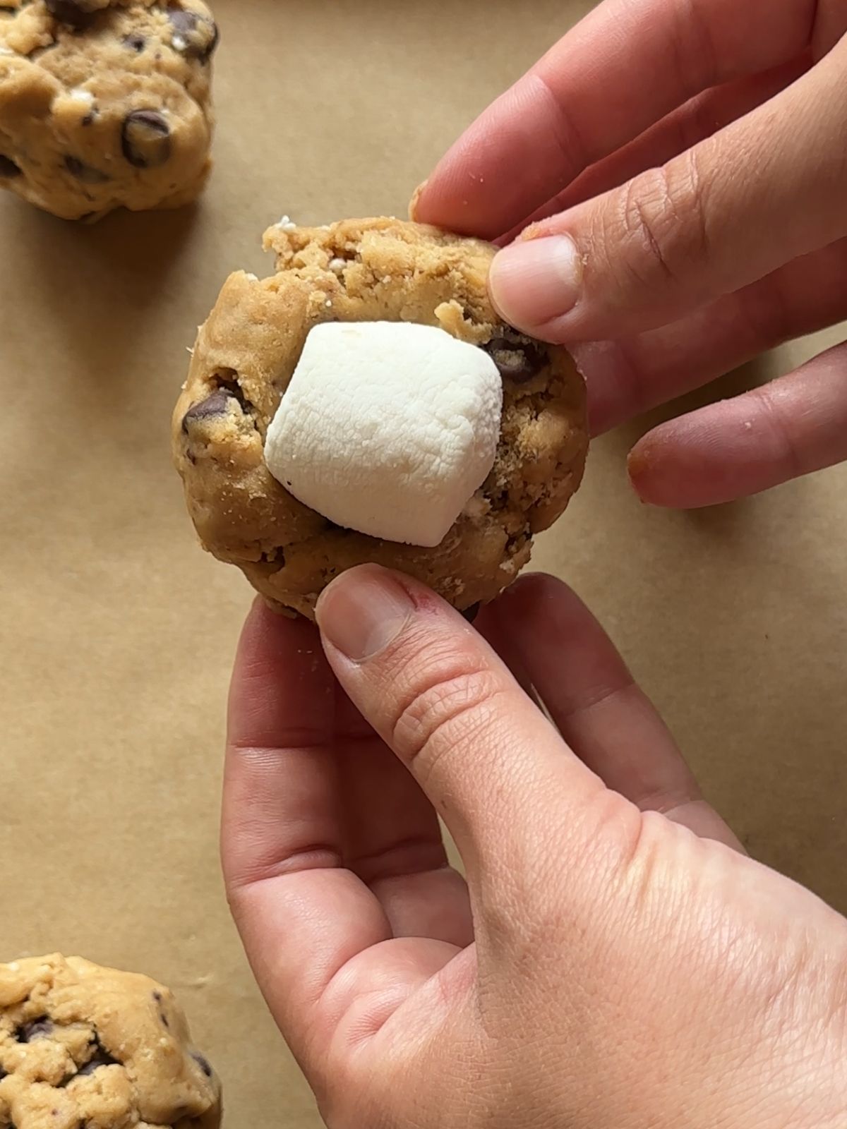 a hand holds a ball of cookie dough with a large marshmallow inserted into the center of the dough