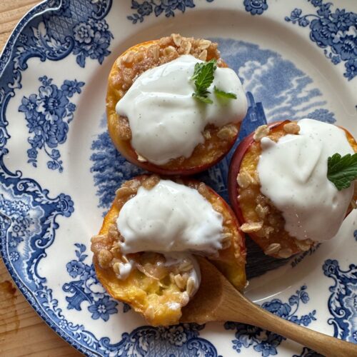 Three sliced and baked peaches are arranged on a patterned blue and white plate. Each peach is topped with an oatmeal crumble and a dollop of vanilla yogurt.