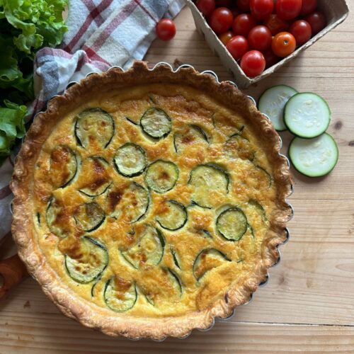 a baked zucchini and onion quiche sits on a wooden countertop, with fresh tomatoes, lettuce, and zucchini slices arranged around it. A checkered towel also lays in the background