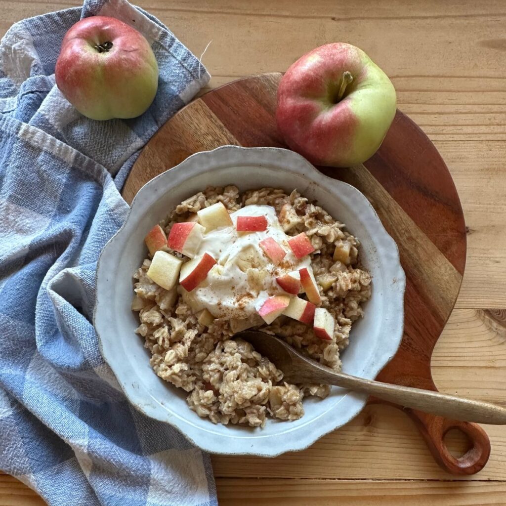 a bowl of apple pie oatmeal, garnished with fresh, chopped apples, a spoonful of vanilla yogurt, and cinnamon. A blue and white checkered towel lays in the background, along with two apples