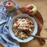a bowl of apple pie oatmeal, garnished with fresh, chopped apples, a spoonful of vanilla yogurt, and cinnamon. A blue and white checkered towel lays in the background, along with two apples