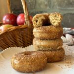 a stack of baked apple cider donuts on a white plate. A basket of red apples is visible in the background