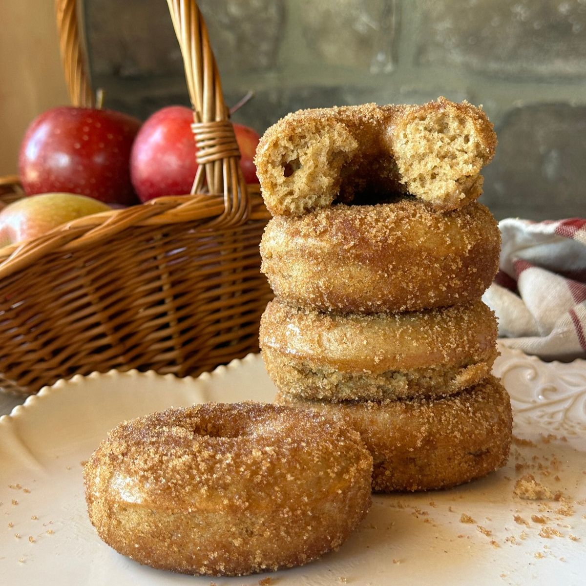 a stack of baked apple cider donuts on a white plate. A basket of red apples is visible in the background